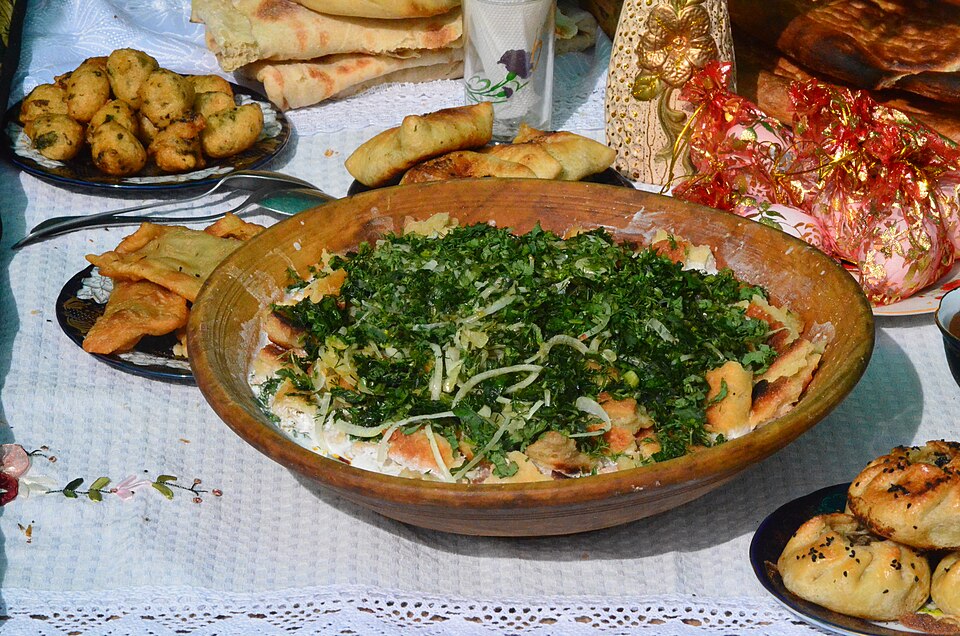 A large wooden bowl of torn flatbread in a pale sauce, generously topped with chopped green herbs and sliced onion.