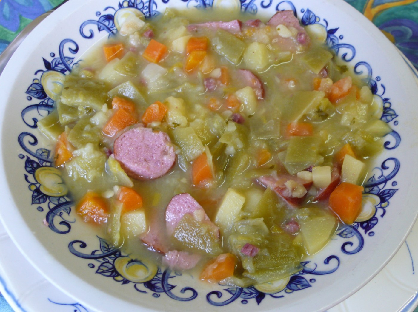 A thick vegetable soup with green beans, potato and carrot pieces, and sliced sausage in a patterned bowl.
